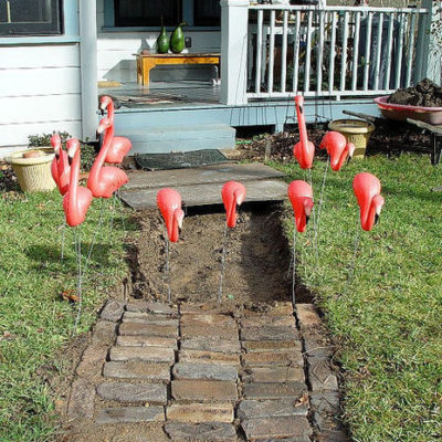 Group of plastic flamingos surrounding hole by stone path and grassy edges.