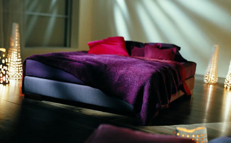 A bed made with a purple cover and red pillow is surrounded by traingle lights set on the wood floor.