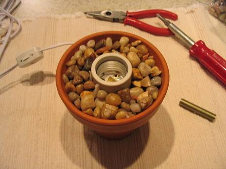 A clay pot full of little rocks surrounding a light socket in the middle is on a beach piece of cloth with a red screwdriver and a pair of red plyers.