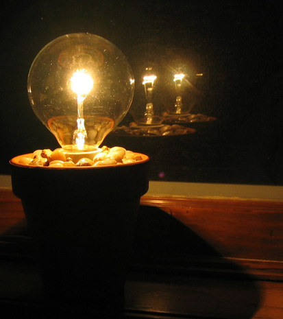 A lit transparent light bulb in a pot with rocks that is reflecting twice in a window.