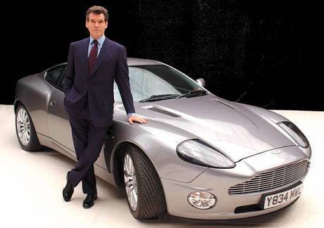 Pierce Brosnan leans on a silver car while wearing a navy blue suit.