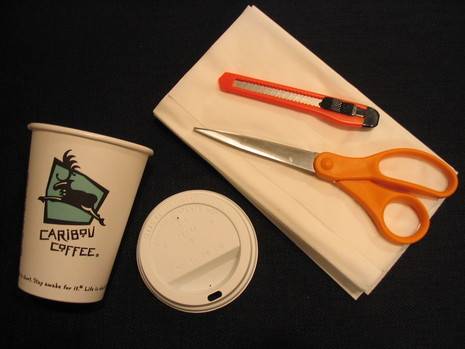 Scissors and another tool on a white napkin near a cup and its lid.