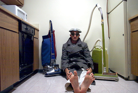 A man sits on the floor of a kitchen between two vaccum cleaners.