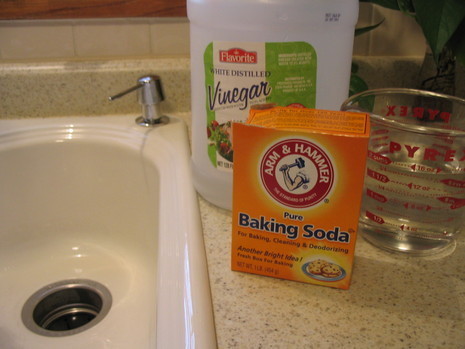 Empty white kitchen sink with baking soda, white vinegar and a measuring cup sitting on the counter.