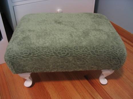 Ottoman upholstered in grayish fabric, with white claw feet on tan hardwood floor.
