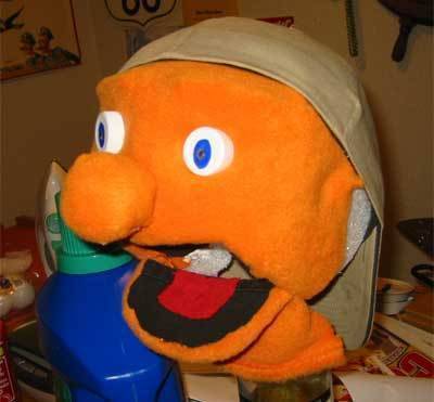 The head of an orange hand puppet with its mouth open, wearing a backwards baseball cap.