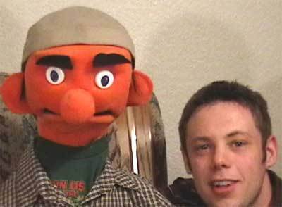 A man is next to a puppet that has an orange face, round nose, and large eyebrows.