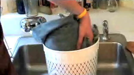 Hand holding a denim fabric item inside a round bin sitting in a stainless steel sink.