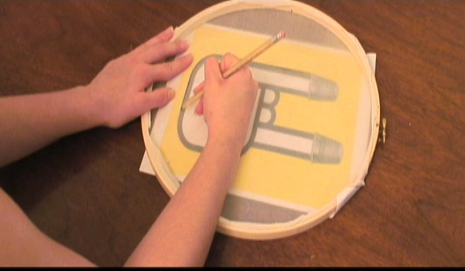 A person uses a pencil to trace an image onto some nylon fabric wrapped around an embroidery hoop.