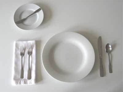 Two silver forks sit on a white napkin next to a white plate with a knife and spoon on the other side, with a smaller plate above them.