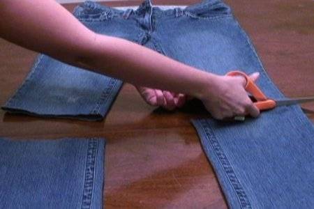 A person cutting the legs of jeans on a table with orange scissors.