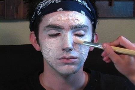 A dark haired man with a bandana on his head sits while someone brushes white makeup onto his face.