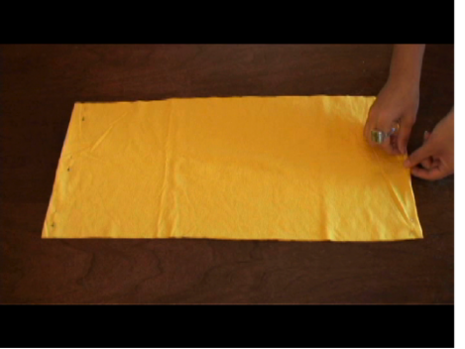 A pair of hands pinching the edge of a piece of rectangular yellow material that is pinned to a piece of wood.