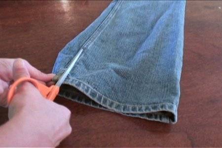A person beginning to cut a pair of blue jeans by the leg seam.