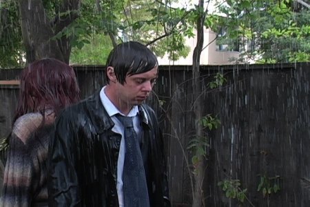 A man and woman standing back to back soaking in the rain outside nearby a wooden fence.