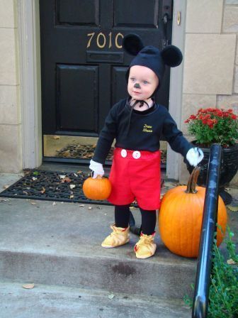 A child on a front porch dressed as Mickey Mouse and standing next to a pumpkin.