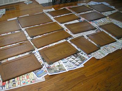 The front pieces of numerous wooden kitchen cabinets lay on newspaper spread out on a hardwoord floor.