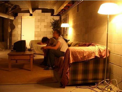 Two men in a basement sitting on a couch with two floor lamps on it sides.