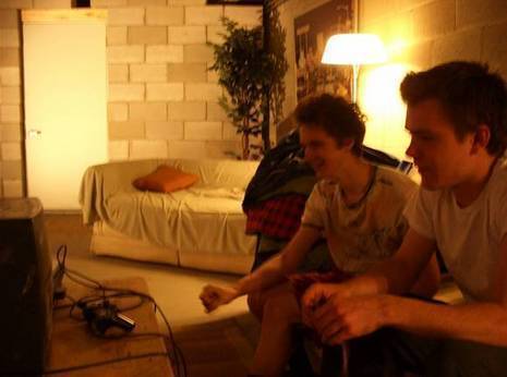 Two young men sit on a couch in an apartment in front of a desktop computer tower.