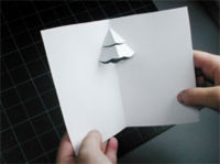 A person holding a piece of paper folded like a greeting card with a partial Christmas tree popped up in the center.