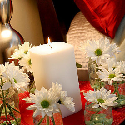 A large white candle surrounded by white jars with white flowers in them, all sitting on a side table next to a bed.