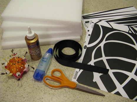 A pin cushion full of pins, a gold tube of glue, a pair of orange handled scissors, four foam boards, some black ribbon and a black and white pattern.