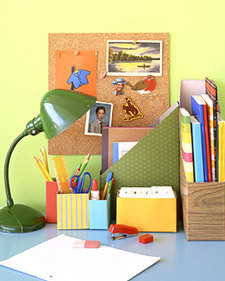 A blue desk with a green lamp, some pencil holders, a magazine holder and a cork board hanging on a yellow wall.