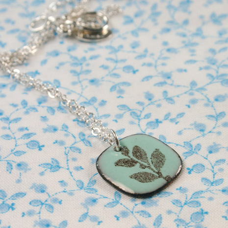 A pendant of a green leaf with a silver chain sits on a blue and white floral cloth.