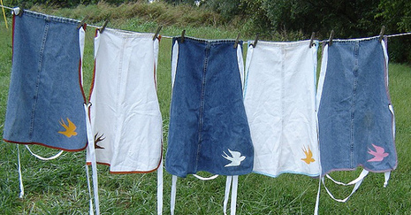 Aprons made of denim are hanging on a clothes line, each a different shade of blue or white.