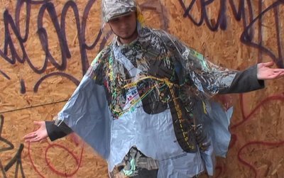 A man with a poncho on standing in front of a wood wall with writing on it.