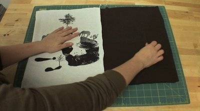 A person's hand holding down a white piece of cloth with a black handprint and horses, and a black piece of cloth, on a green cutting mat.