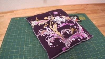 A purple pillow with a pink feather design is laying on a green cutting mat.