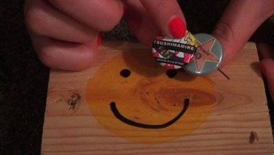 A person with red nail polish holds two pins over a wooden square with a smiley face painted on it.