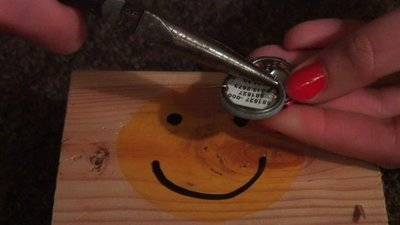 A hand with red nail polish is holding a piece of metal over a block of wood with a yellow happy face on it.
