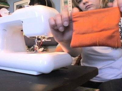 Person sitting behind a white sewing machine and holding up a square of orange fabric.