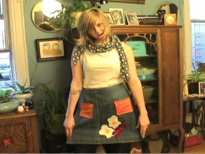 A woman wearing a jean skirt with bright patches on it smiles at the camera.
