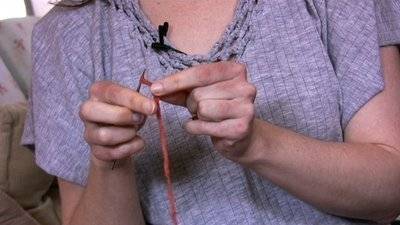 A woman in a gray shirt is holding a knitting needle in one hand with thin yarn of heavy string wrapped around the needle and pinched between two fingers to hold it in place.