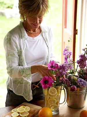 An older woman standing at a table with a series of pots and flowers in front of her.