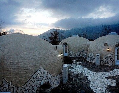 Several round concrete homes are arranged around a white brick path, with mountains in the background.