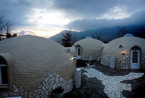 Several round concrete homes are arranged around a white brick path, with mountains in the background.
