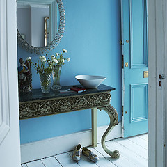 A cornflower blue room with white wood floors and a fancy endtable and mirror.