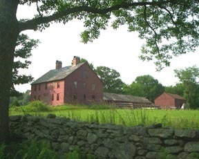 A two story red house with a small red barn beyond a rock wall and green field.