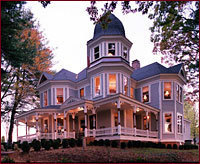 A two story victorian house with a wrap around porch and a widow peak that is lit at dusk.