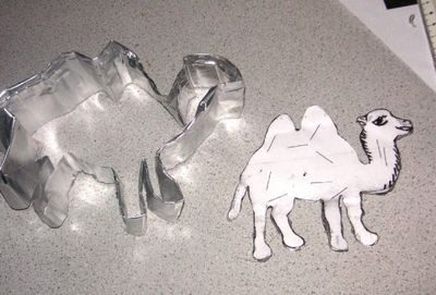 A cookie cutter in the shape of a camel lays next to a camel made of cut paper.