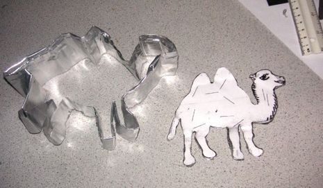A cookie cutter in the shape of a camel lays next to a camel made of cut paper.