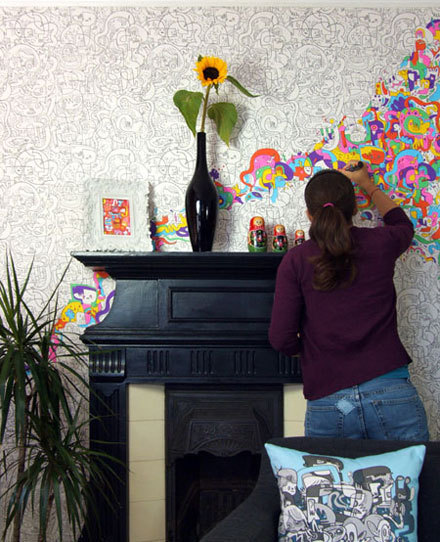A woman painting her wallpaper in splashes of neon color, next to a tall black mantle and a vase with a yellow sunflower.