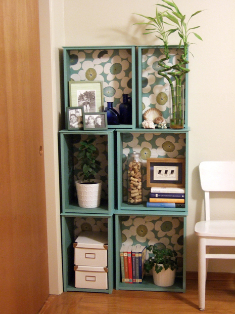 A blue/green six section shelve housing three plants, some pictures, some books and shoe boxes is standing next to a white chair.