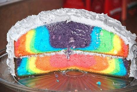 A two-layer round cake has a rainbow interior and white frosting.