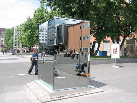 A toilet cubicle with mirrored walls sits in a very public space.