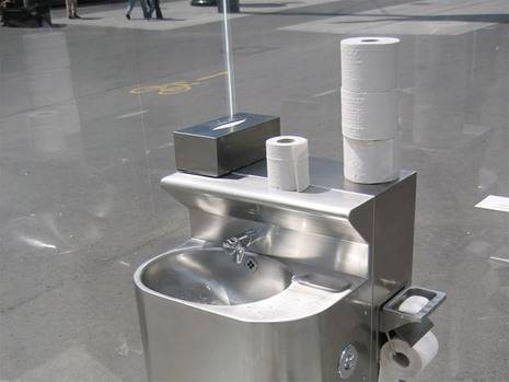 A silver mobile sink sits in an outdoor area with toilet paper, paper towels, and Kleenex.